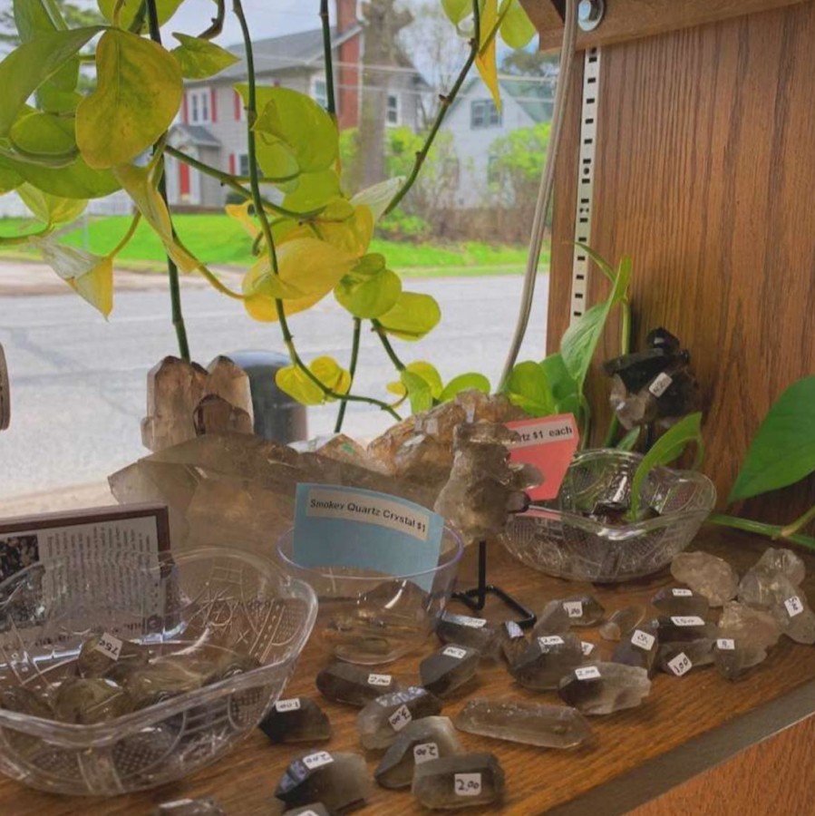 The photo displayed represents a small corner of the shop and some of its belongings. Shown are several pieces of smokey quartz, selenite, and sunstone with several plants and windows to enhance the environment. 