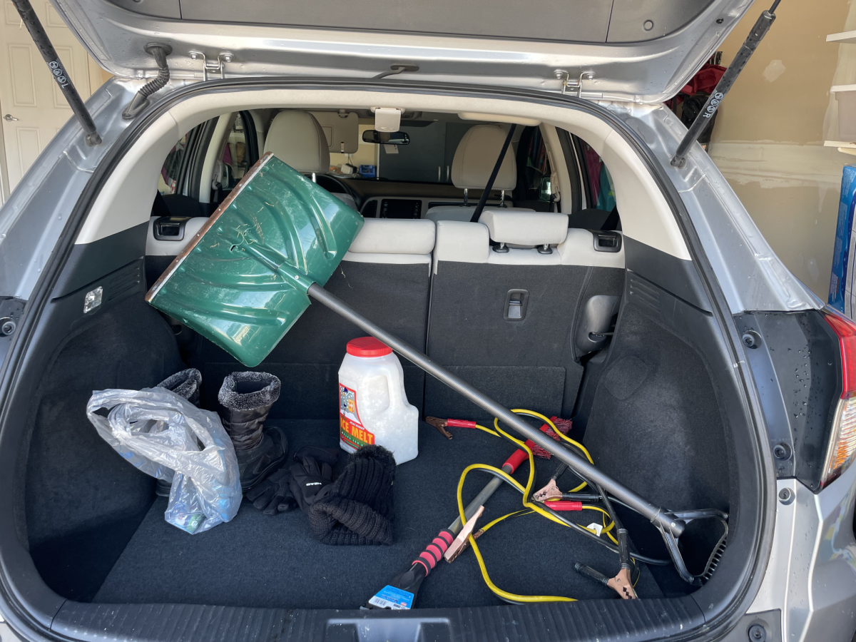 Car trunk loaded with supplies including jumper cables, shovel, winter gear, food, flashlight, salt and ice scraper.